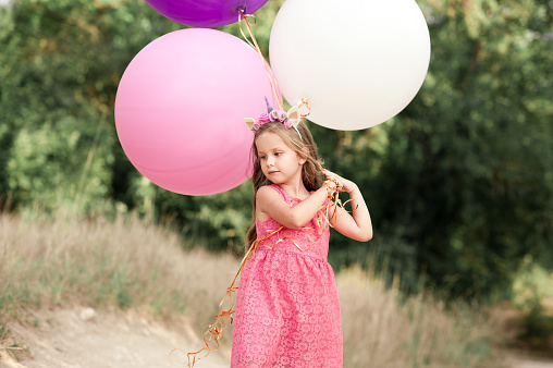 Cute kid girl 4-5 year old holding colorful balloons over green nature background. Playing outdoors. Childhood.