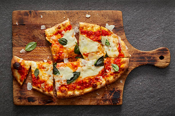 A sliced neapolitan pizza on a rustic wood board placed on a black slate stone surface.  The pizza is cut with missing pieces and parmesan cheese sprinkled on top.  Please see my portfolio for other food and drink images. 