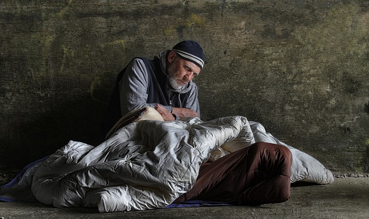 Bearded Homeless Man in His 40s Looking at Camera While Sitting Under the Bridge. Homelessness Social Issues Concept.