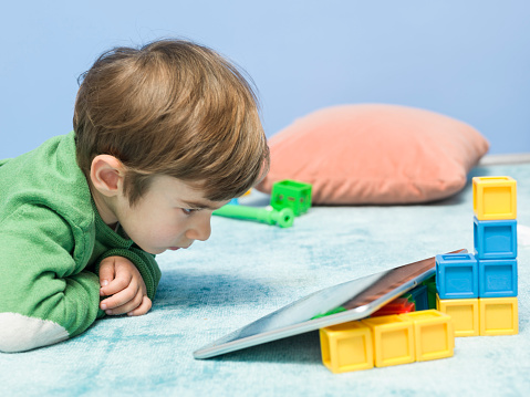 Little boy sitting on blue carpet and watching film on tablet pc.He has blonde hair and wearing green sweatshirt. Various toy blocks in various colors are on carpet.Few cushions are seen around.Shot in studio with medium format DSLR camera Hasselblad.