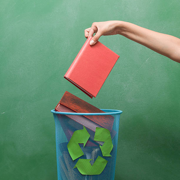 Human hand holding a red covered book over garbage bin.The bin is full of books and has a recycling sign on it.The bin is on the down left of frame while female human hand is on the right side of frame.The image was shot in studio with medium format DSLR camera Hasselblad, cropped to square compostion