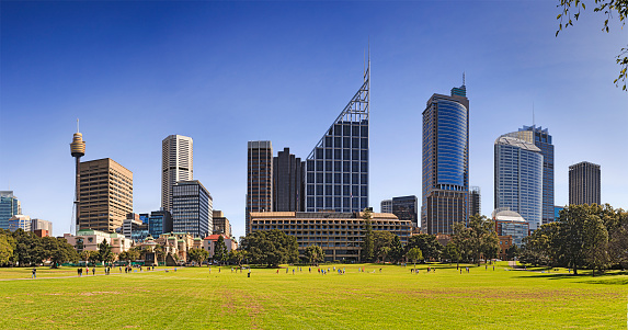 Wide panorama of Sydney city landmark towers and buildings from Royal eye hospital to modern bank offices as seen across green grass of domain park on a bright sunny day.