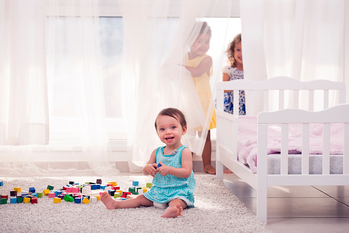 Three children playing together in playroom. Baby girl sitting on the carpet playing with colorful block cubes, two little girls in age 2.5 years old playing hide and seek, hiding behind the curtain.