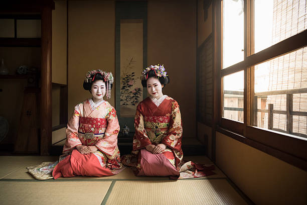 Two Japanese maiko kneeling on floor wearing kimonos Young women in traditional maiko costume sitting on floor in Japanese building interior geisha costume stock pictures, royalty-free photos & images