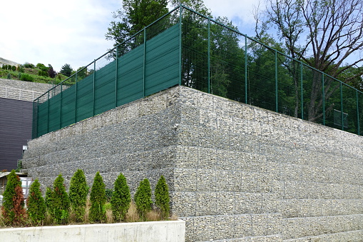 Protective Gabion Wall Filled With Raw Rocks, Stones Or Concrete Blocks And Tied With Thick Metal Wire On Landscape With Blue Sky And Trees Near Living Quarters