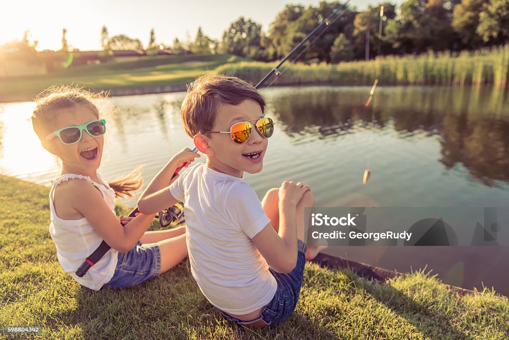 Kids catching fish Funny stylish little boy and girl in sun glasses are looking at camera and smiling while catching fish in the pond using a fishing rod, sitting on the ground Child Stock Photo
