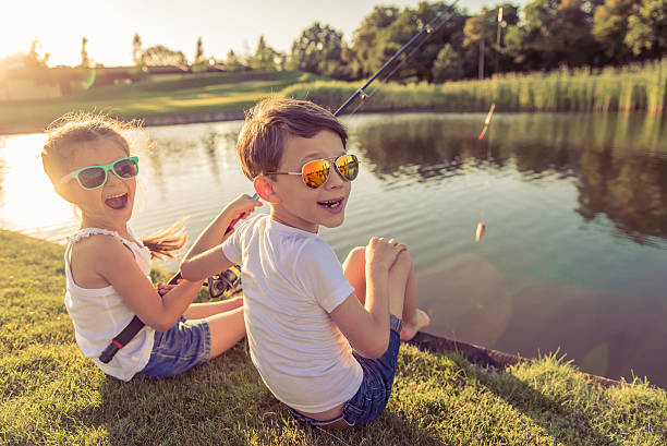 Kids catching fish Funny stylish little boy and girl in sun glasses are looking at camera and smiling while catching fish in the pond using a fishing rod, sitting on the ground catching photos stock pictures, royalty-free photos & images