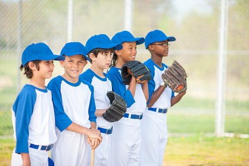 A multi-ethnic group of elementary age boys are standing in a row before their baseball game. They are wearing their baseball uniforms, baseball hats, and mits.