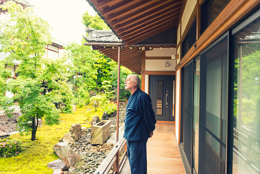 Senior man in dark blue kimono standing on wooden porch outside of a temple building and observing temple garden. He stand against large screen doors. In garden are green trees, moss and stones. Japan.