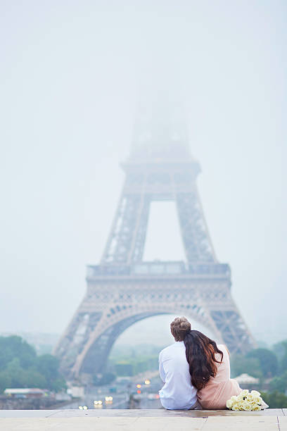 Romantic couple together in Paris Beautiful romantic couple in love with bunch of white roses sitting near the Eiffel tower in Paris on a cloudy and foggy rainy day, back view paris france eiffel tower love kissing stock pictures, royalty-free photos & images