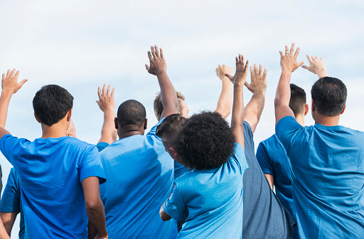 Rear view of a group of multi-ethnic men and boys wearing blue shirts, raising their hands. They are a group of volunteers working on a community service project.