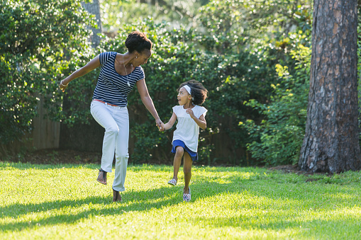 An African American mother with her beautiful young mixed race daughter, holding hands and skipping as they smiling at each other. It is a sunny day in the park or back yard on the grass, with trees and foliage in the background.