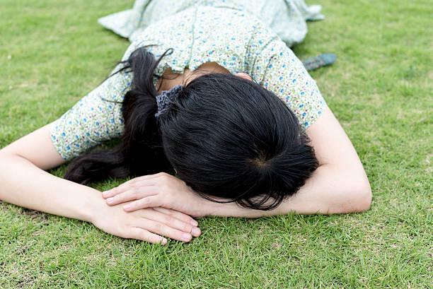 Young joyful woman lies on green grass. Happy girl is relaxing at chamomile field in warm summer day on background of big trees in park.