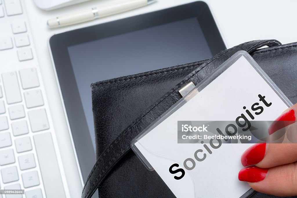 Woman showing her occupation on a name tag Mature Woman showing Sociologist occupation on a name tag  on her bag with tablet and  pc keyboard Adult Stock Photo