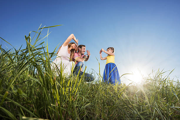 A small family of four lay in the grass together on a sunny summer day as they pose for a portrait after a workout.  They are each dressed comfortably in athletic wear and are smiling.