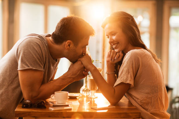 Young romantic man kissing girlfriend's hand in a cafe. Smiling woman enjoying in a cafe while being kissed in a hand by her boyfriend. heterosexual couple stock pictures, royalty-free photos & images