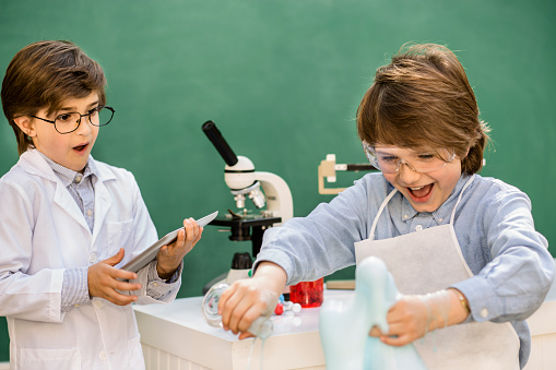 Two excited elementary-age children collaborate on various science experiments.  They are using chemical glassware, chemicals as their experiment is causing a chemical reaction.  Microscope on table.  They wear safety and eyeglasses.   Imagination, creativity, inspiration for these little chemists!  Chalkboard, classroom setting.