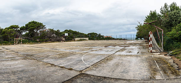 Old abandoned school sports court or schoolyard for different ac Old abandoned school sports court or schoolyard for different activities. Ruins of a sport venue abandoned long time ago with soccer, handball or football goals, basketball hoops and boards and destroyed concrete plates. tennis ball container stock pictures, royalty-free photos & images