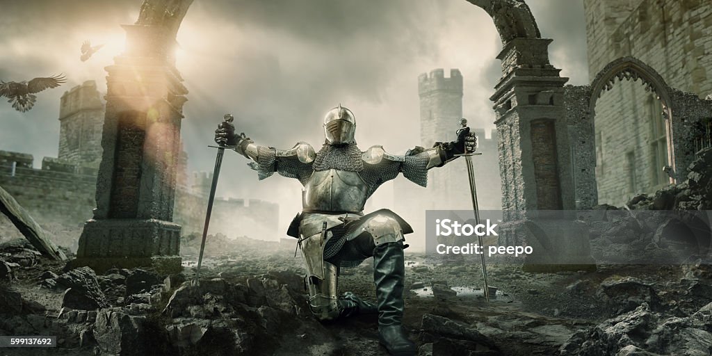 Medieval Knight Kneeling With Sword In Front of Building Ruin A medieval knight in a suit of armour, kneeling on knee holding up a sword in each outstretched hand. The knight is alone in front of a ruined medieval arch and outbuildings in front of a castle, surround by rocks and rubble under a dramatic stormy evening sky. Knight - Person Stock Photo