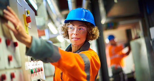 boiler room engineer A female industrial service engineer conducts a safety check of a control panel in a boiler room. She is wearing hi vis, hard hat, safety glasses and holding a digital tablet as she conducts a safety inspection. She is reaching for one of the dials as she looks to camera. power plant workers stock pictures, royalty-free photos & images