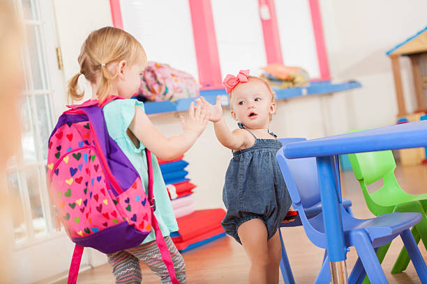 Cute blonde Caucasian sisters, friends say hello and high five in their daycare classroom. The girls are standing in the classroom and ready for the day.