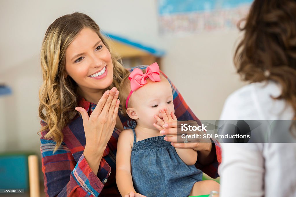 Smiling mother and her cute toddler daughter practicing sign language Sweet little toddler girl with pink hair bow is sitting on smiling Caucasian mother's lap. Mother is using "thank you" sign language with daughter and preschool teacher. Sign Language Stock Photo