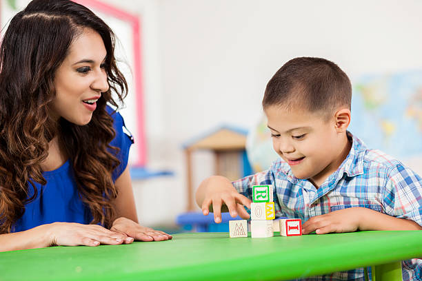 Down Syndrome boy playing with building blocks at daycare Happy Hispanic Down Syndrome boy is sitting at a table with his female Hispanic teacher and playing with building blocks. They are sitting at a table and playing in the learning center of his daycare. special education stock pictures, royalty-free photos & images