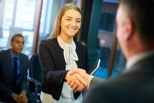 young woman steps forward as she is called in for her interview and shakes hands with her possible new employer. Behind her are some other candidates waiting for their turn .