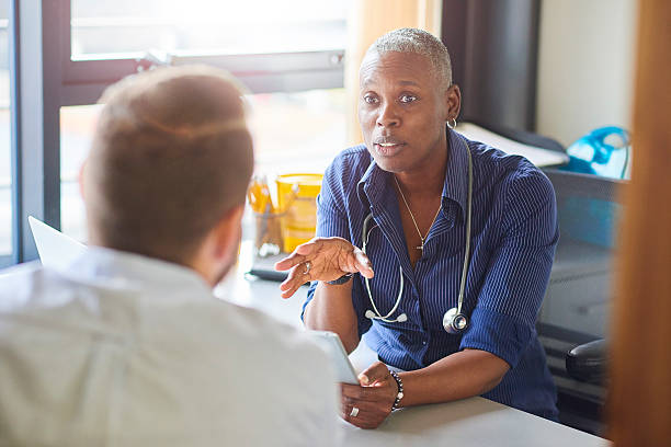 Doctor chatting to male patient A female doctor sits at her desk and chats to a male patient while looking at his test results on her digital tablet .  She is a blue shirt with the sleeves rolled up and a stethoscope around her neck. general practitioner stock pictures, royalty-free photos & images