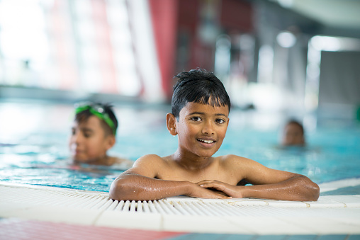 A multi-ethnic group of elementary age children are taking swimming lessons at the pool. A young boy is at the edge of the pool and is smiling while looking at the camera.