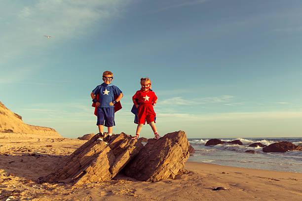 Two young children a boy and a girl are dressed up in superhero capes and masks stand on a rock on a beach in California, USA. They stand with hands on their hips while they look at the camera.  