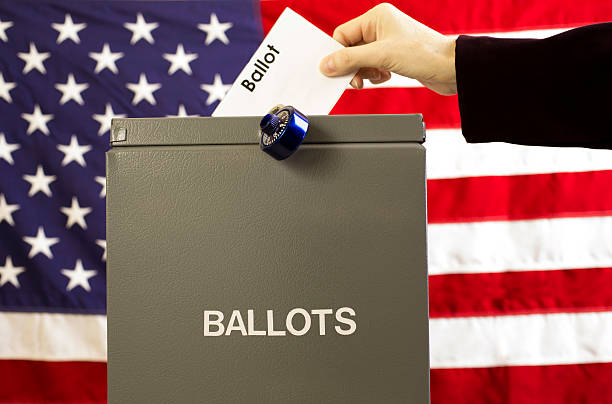 istock Voter inserting ballot into box with USA flag background. 599463128