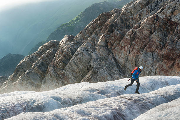 Male hiker wearing crampons, backpack, helmet and jacket jumps across an icy crevasse while trekking on a solo adventure on Lemon Glacier with granite mountains and temperate rainforest in background, Juneau Icefield, Juneau, Alaska, USA