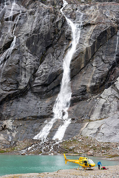 Alaska, USA - August 6, 2016: Adult male unpacks gear from a yellow helicopter that landed on the shore of a mineral lake with a giant waterfall to drop off passengers for an overnight trekking adventure on Lemon Glacier, Juneau Icefield, Juneau