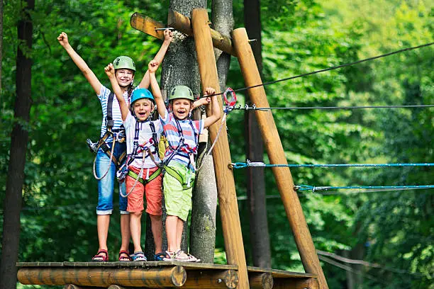 Little girl aged 10 with her brothers aged 7, wearing helmets stadning on wooden platform holding zip line in the outdoors ropes course adventure park. Kids are smiling at the camera and cheering.