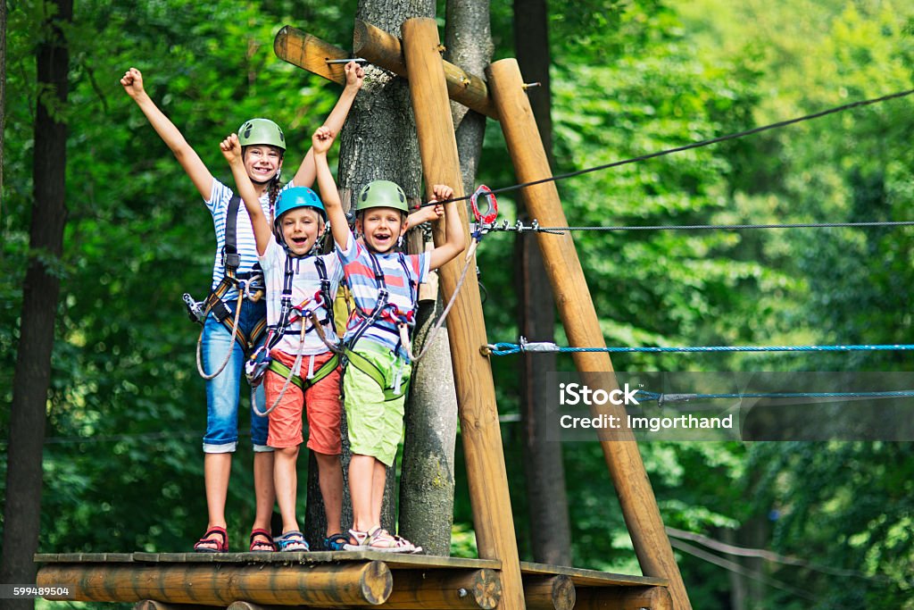 Kids having fun in ropes course adventure park Little girl aged 10 with her brothers aged 7, wearing helmets stadning on wooden platform holding zip line in the outdoors ropes course adventure park. Kids are smiling at the camera and cheering. Child Stock Photo