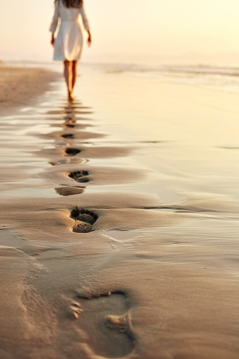 Rear view of woman walking barefoot in distance on wet sand. Female in sundress is leaving her footprints on shore. Low section of woman spending leisure time on beach at sunset.