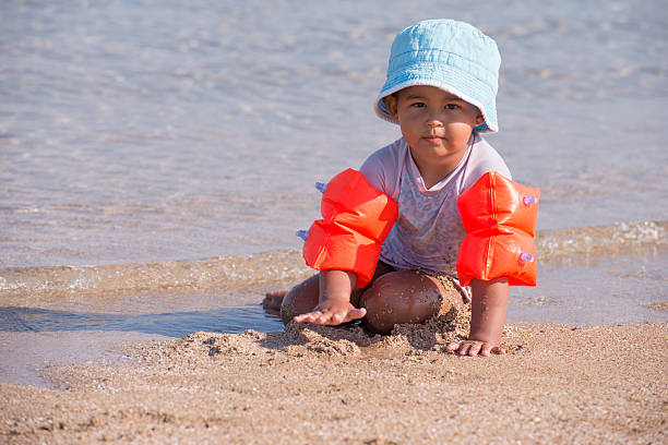 Child wearing inflatable armbands / water wings / swimmies / floaties, playing with sea sand, sitting on water's edge, wearing swimsuit and hat protect against the sun UV.
