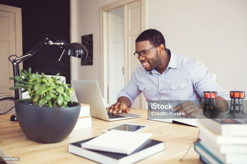 Afro american young man in a home office Afro american young man sitting at the desk in a home office, using laptop and smart phone. Working At Home Stock Photo