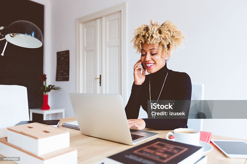 Afro american young woman in a home office Beautiful afro american young woman sitting at the desk in a home office, using laptop and talking on mobile phone. Adult Stock Photo