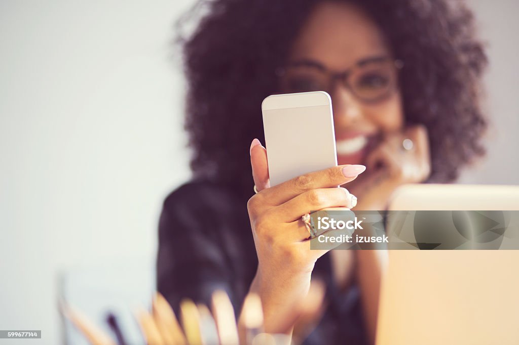 Beautiful woman using smart phone at home Beautiful afro american young woman sitting at the desk and using a smart phone. Focus on mobile. Adult Stock Photo