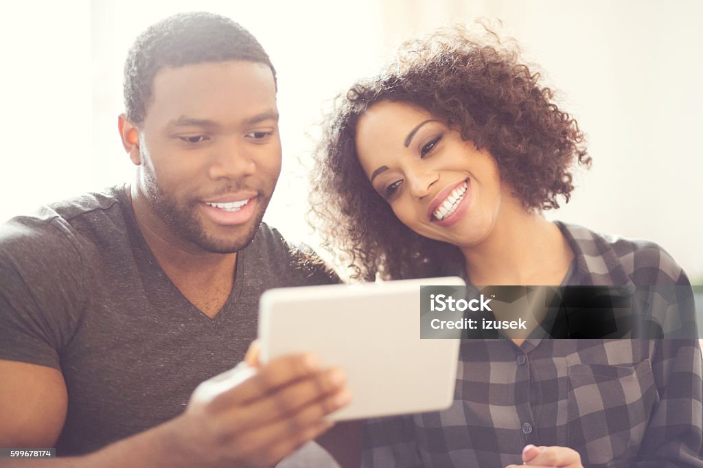 Happy afro american couple using a digital tablet at home Afro american friendly couple sitting on sofa at home and using a digital tablet. Focus on faces. Adult Stock Photo