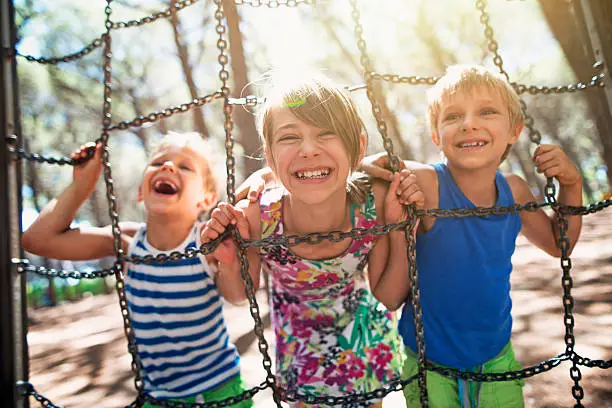 Three kids having fun at the playground. The girl aged 10 and boys aged 7 are laughing at the camera. Sunny summer day. Kids are holding climbing chains at the playground.