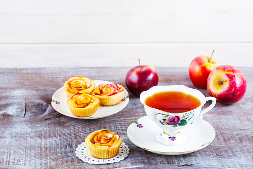 Cup of tea and apple roses shaped muffins on rustic wooden table. Sweet apple dessert pie. Homemade apple rose pastry. Breakfast tea with sweet apple pastry