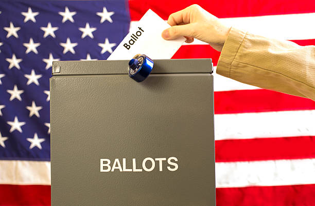 istock Voter inserting ballot into box with USA flag background. 599748944