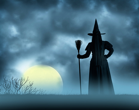 A Halloween witch holds her broom in an upright position as she stands in an open field and watches a full moon rise over the horizon.