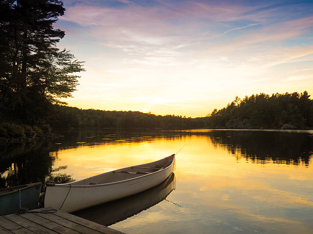 This is a square, color, royalty free stock photograph of a scenic lake landscape in New York State's Catskill Mountains. A simple, wooden dock extends into the reflective water's surface. A classic rowboat is docked near the shore. Green summer trees fill the background and line the edges of the country scene. Photographed with a Samsung Galaxy S5 mobile phone.