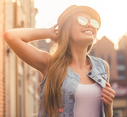 Beautiful stylish young girl in casual clothes, hat and sun glasses is looking at the sun and smiling while walking outdoors
