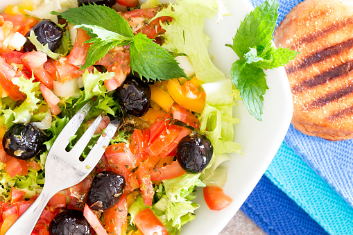 Fresh Mediterranean salad with lettuce, tomato, sweet peppers and black olives served with crispy toasted bread on blue napkins, close up overhead view