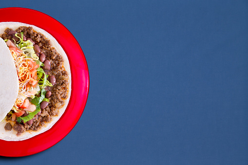 Soft taco with savory filling and vegetables served on a red plate over a blue background with copyspace, overhead view